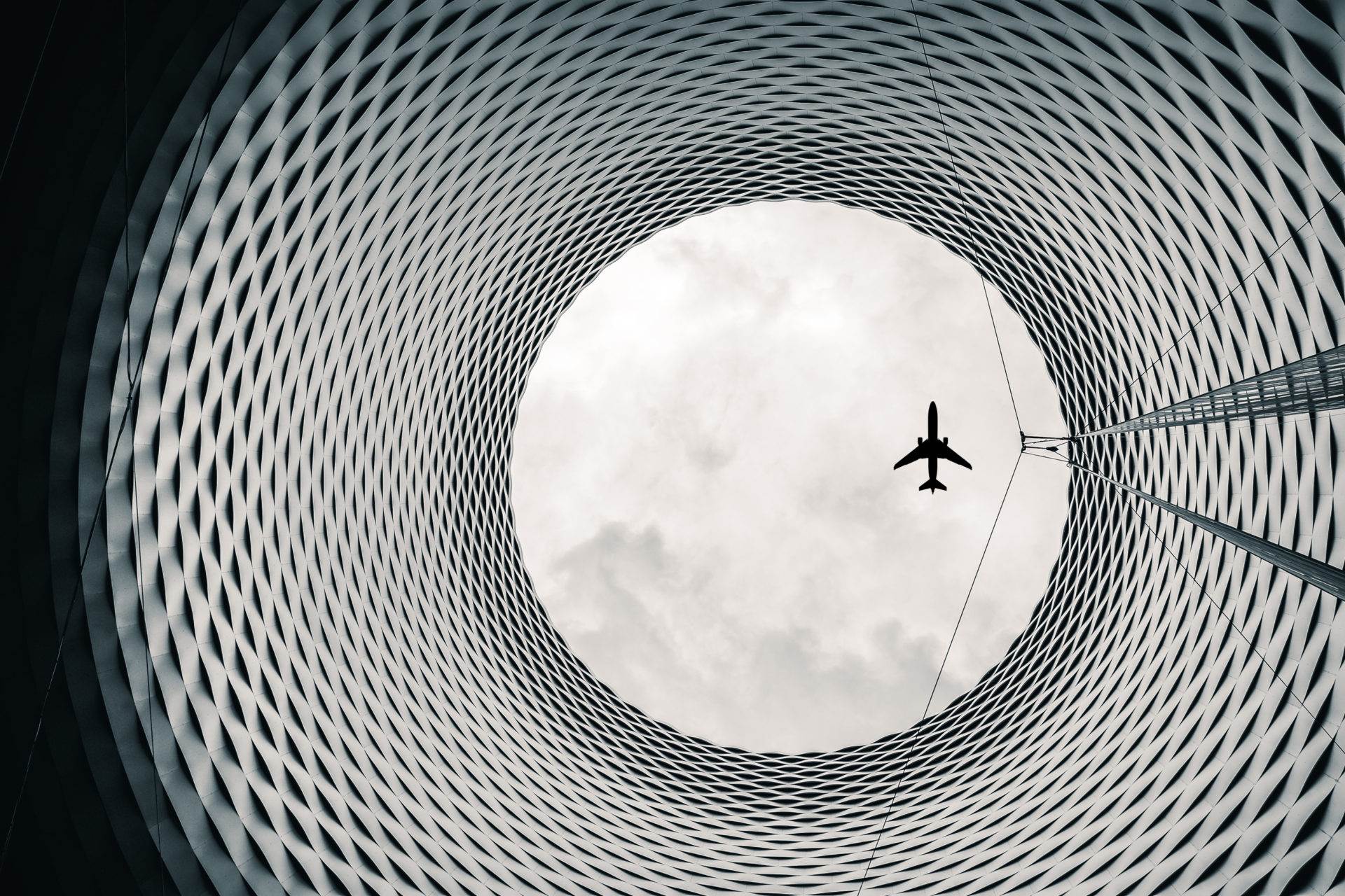 Architekturfotografie: Blick durch eine runde, geflochtene Fassadenöffnung in den Himmel mit Flugzeug